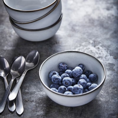A bowl of blueberries in a small white stoneware bowl next to a stack of similar bowls and silver spoons.
