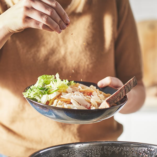 Person seasoning noodles with cheese and veggies on a plate.