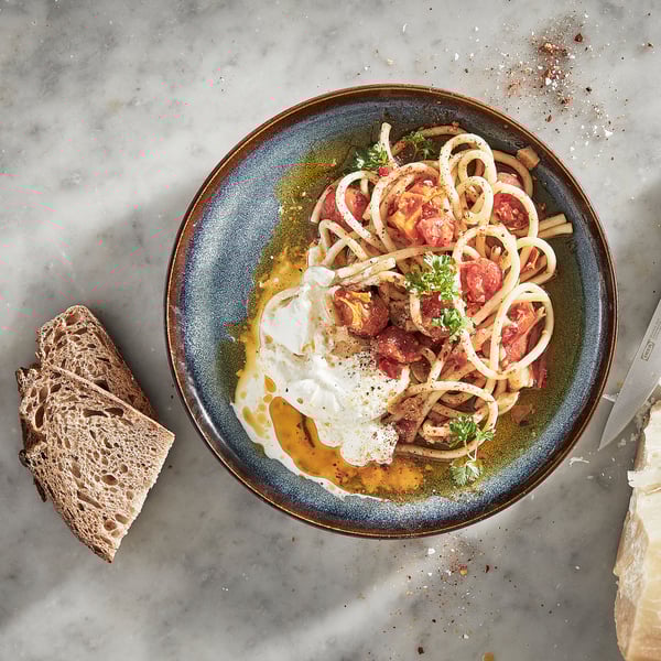 A blue glazed ceramic plate with spaghetti, burrata, and tomatoes, accompanied by rye bread and knife on a marble surface.