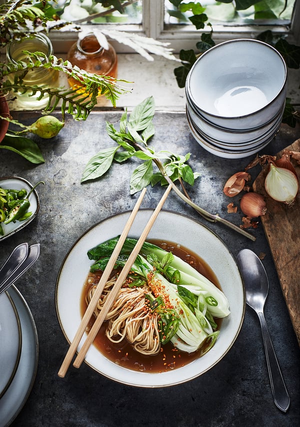 A bowl of noodle soup sits on a rustic table with chopsticks, surrounded by vegetables and stacked white plates.