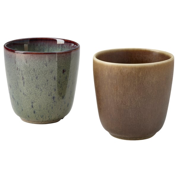 Two ceramic cups, one green with dark spots, one brown, rustic, handcrafted design, rested on a white surface.