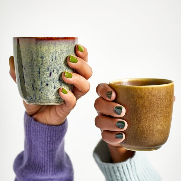 Two hands hold mugs with green nails; one mug greenish-blue, other mustard.