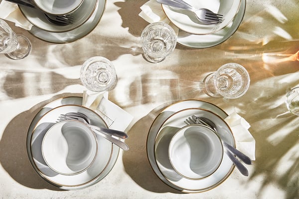 Four elegant, white, sandy-glazed GLADELIG plates with gold rims set with silverware and glasses on a sunlit table.