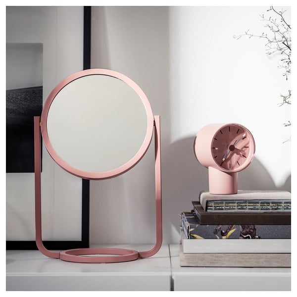 Pink GRANVÅG table mirror, round with a metal base, positioned on a shelf with a stack of books and a pink clock.