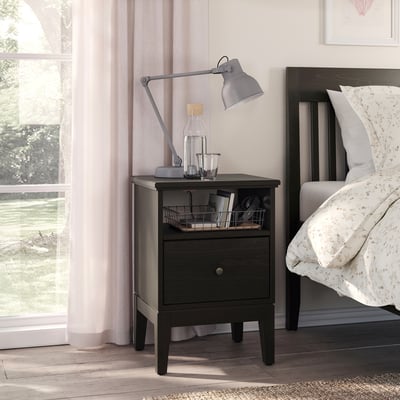 Bedroom with black ikea nightstand, lamp, and jars beside a white bed and window with pink curtains.