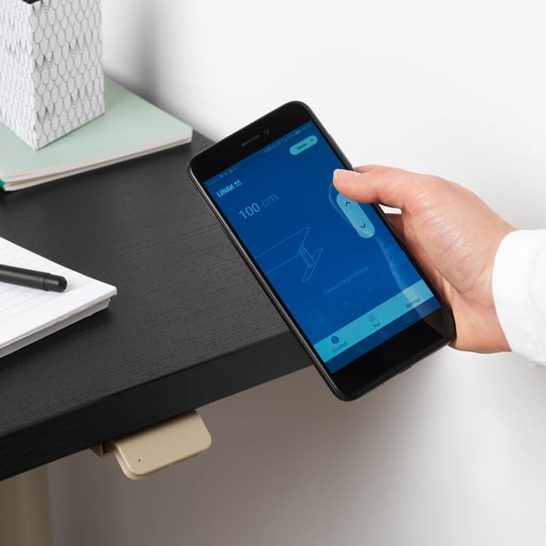 Person adjusts desk height using smartphone app. IDÅSEN desk with remote control and height adjustment from 63 to 127 cm.