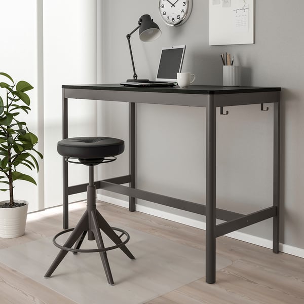 Modern black metal desk with sleek, square top & round stool. Neatly fits against grey wall, larger than plant nearby.