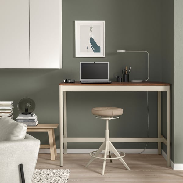 Minimalist workspace with IDÅSEN white table. Laptop, lamp, supplies, and simple stool. Green wall and stackable books at side.