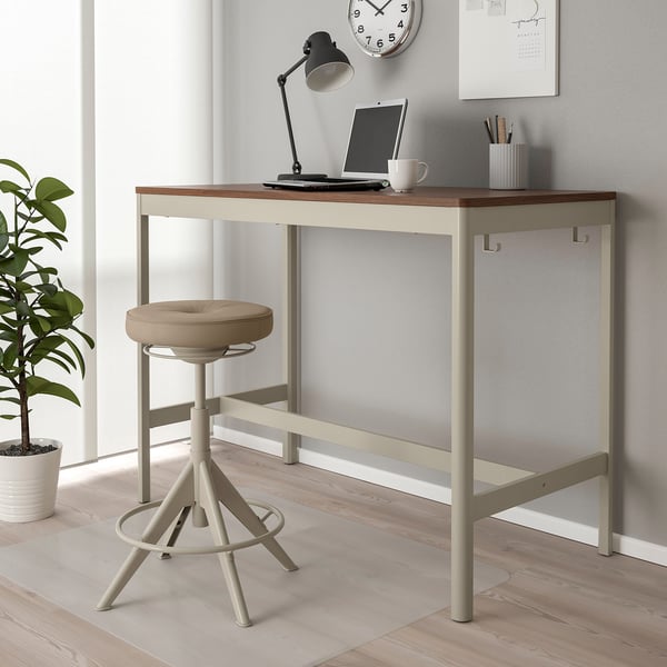Modern IDÅSEN desk setup. Light wood top, beige metal legs. Clock, lamp, laptop, and plant beside.