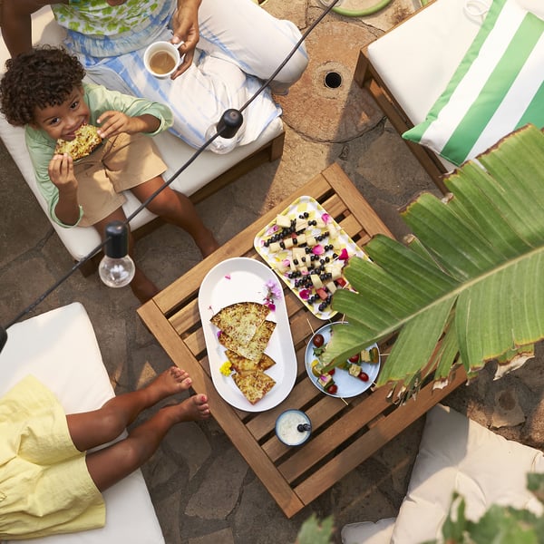 Family enjoying brunch on patio. Children eat pizza, adults sip coffee. Wooden table holds plates with food, including pizza and fruit, and candles.