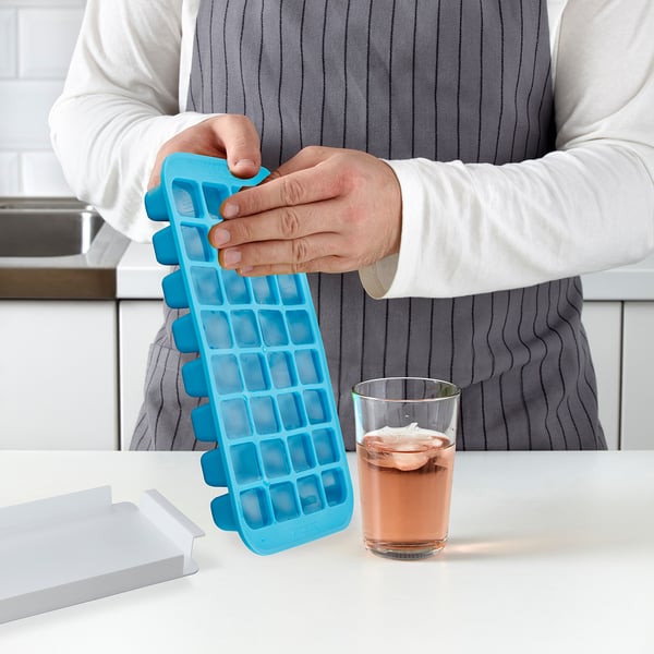 Person holding blue ice cube tray with 32 compartments, showing its easy push-out feature. Glass of drink with ice beside it.