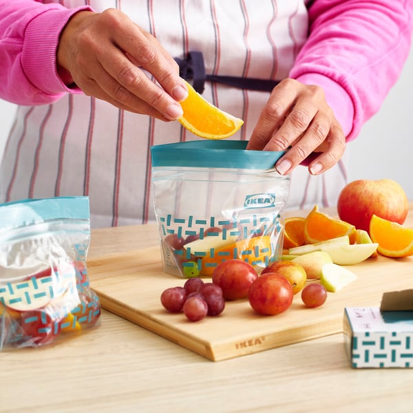 A person puts orange slice into an ikea reusable storage bag filled with fruits on a wooden board.