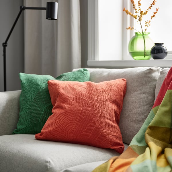 Red-orange leaf-patterned JÄTTEGRAN cushion on grey couch with green pillows and multicoloured throw.