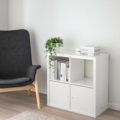 White ikea KALLAX shelving unit with books and plant, next to large grey chair on light wooden floor.