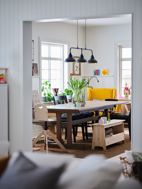 Bright dining area with wooden table, black chairs, yellow bench, and three black pendant lights hanging above, featuring a cosy and colourful design.