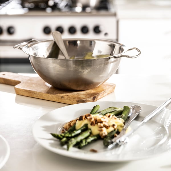 Silver bowl with handled lid on wooden stand. Plate with green food and sauce in foreground.