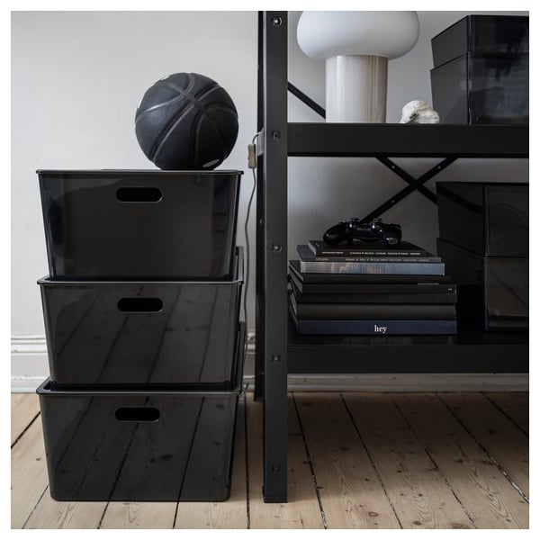 Black plastic storage boxes stacked on floor, with basketball on top, next to metal shelf with games and decor.