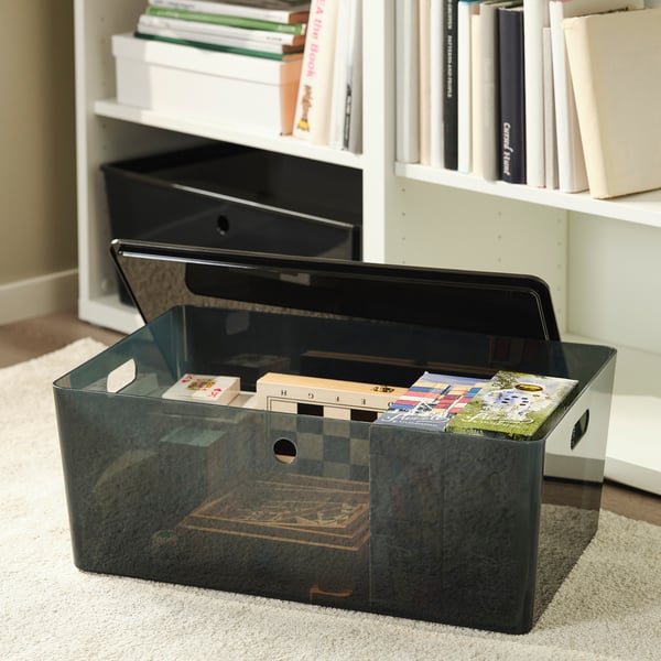 Open black plastic storage box on carpet, holding board games, in front of bookshelf. Handles on sides for easy carrying.