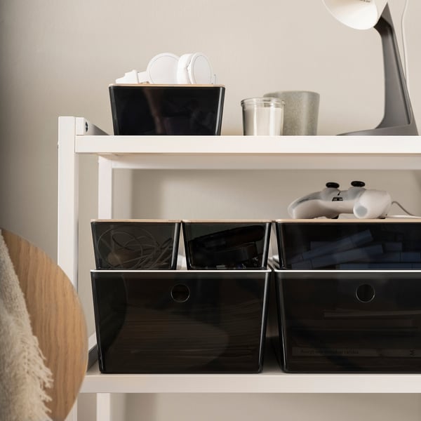 Organised shelf with KUGGIS storage boxes in dark grey, containing cables and tech accessories, fitting neatly under a game controller.