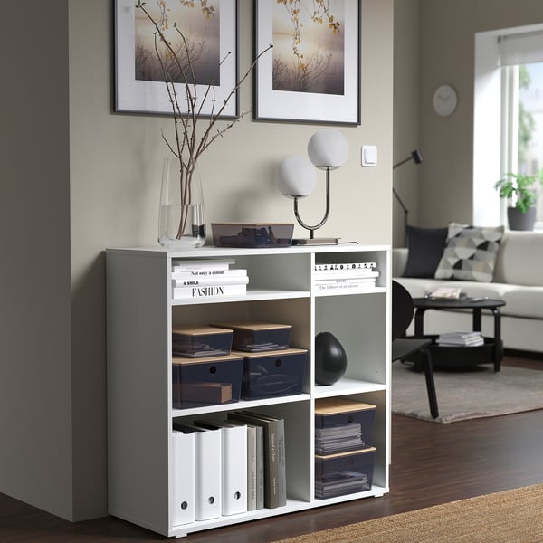 White KUGGIS storage box with blue bins, books, and lamp on a shelf. Its compact size fits neatly under framed pictures.