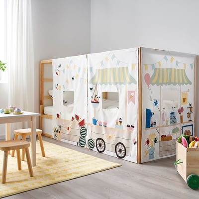 Childrens room with white KURA bunk bed, decorated with colourful market tent. Yellow rug, wooden chairs, and toys are arranged around.