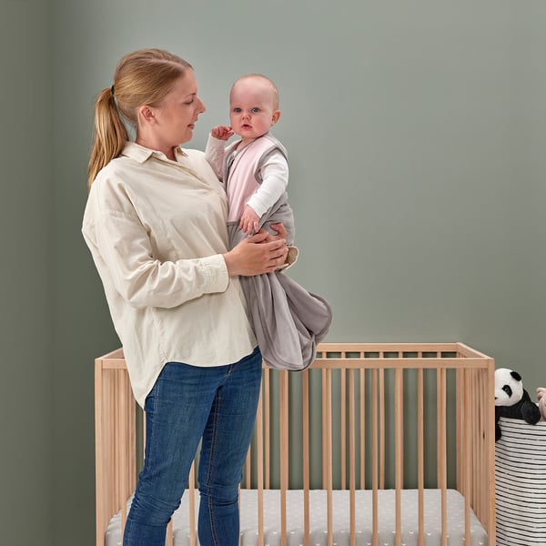 Person holds baby in pink sleep bag by safe crib with toys.