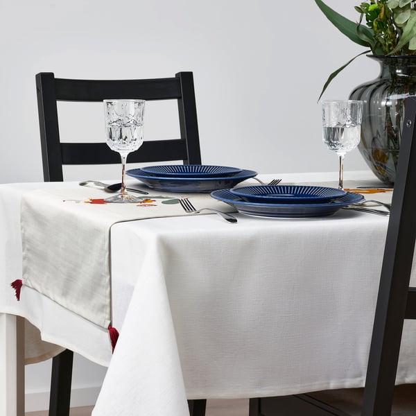 Dining table set with blue plates, glasses, silverware, on white MÄVINN runner, flanked by black chairs.