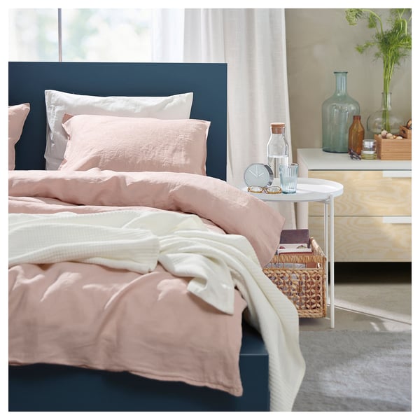 A cosy modern minimalist bedroom with a light pink bed, dark blue headboard, white bedside table with a clock, vase of flowers, and a basket of books.