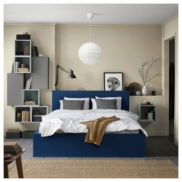Modern bedroom with blue MALM bed, white linens, grey pillows, wall shelves with books, large white pendant light, and a floor lamp.