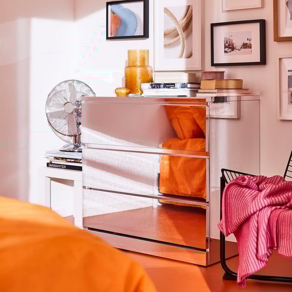 Mirrored pink MALM chest, three drawers, reflecting wall art. Fan and books on top, orange throw over side.