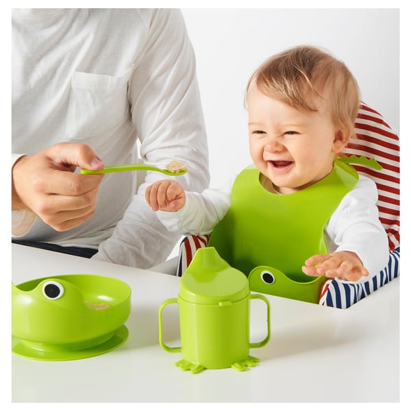 Baby eating with vibrant frog-themed feeding set. Adult spoon-feeds with green utensils, bib, and cup on a table.