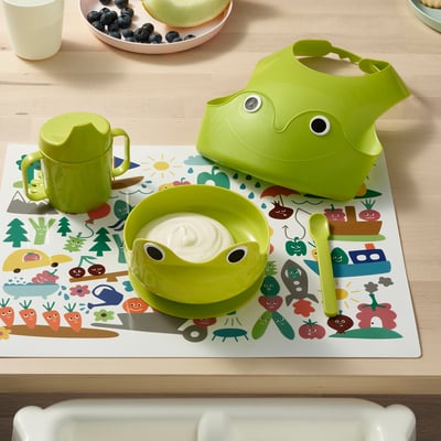Childs mealtime set: green frog bowl, cup, spoon, and bib on a colourful placemat.