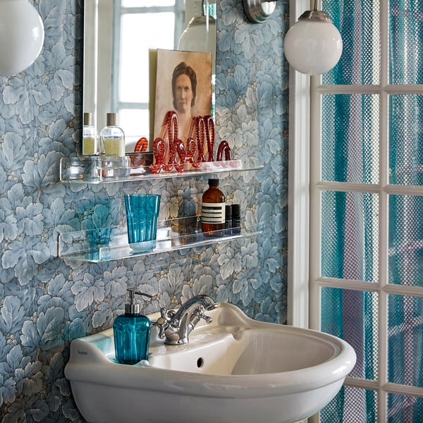 Bathroom with blue floral wallpaper, white sink, clear glass shelves holding various toiletries and a framed photo.