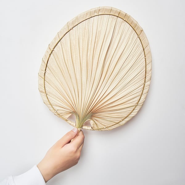 Person holding round, woven palm leaf fan against white wall.