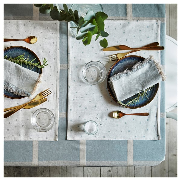NISSÖGA natural cotton-jute tablecloth, blue-white dishes, gold cutlery, glasses on wooden table with greenery.