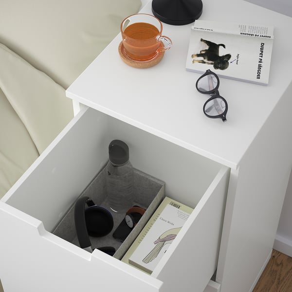 White bedside drawer open, showing a water bottle, clock, and books. Top has tea, book, and glasses.