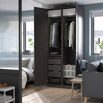 Modern bedroom with open dark PAX wardrobe showing clothes, drawers, and storage bins beside a grey couch and white shoe rack.