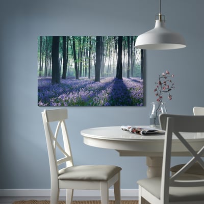 Dining room with PJÄTTERYD forest painting on light blue wall. White chairs around table with pink flowers and pendant lamp.