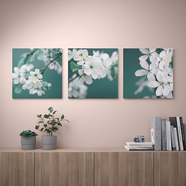 Three PJÄTTERYD prints with white flowers on green backgrounds hang on a pink wall above a wooden shelf with potted plants and books.