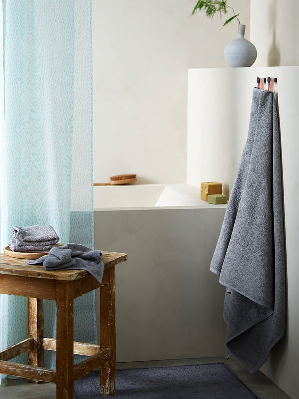 Modern bathroom with grey marble sink, RÅNEÄLVEN turquoise shower curtain, wooden stool with grey towels, hanging grey towel, brushes, and plant.