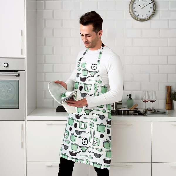 Person in kitchen wearing a patterned apron, holding a white cloth, near an oven and counter with glasses and dishes.