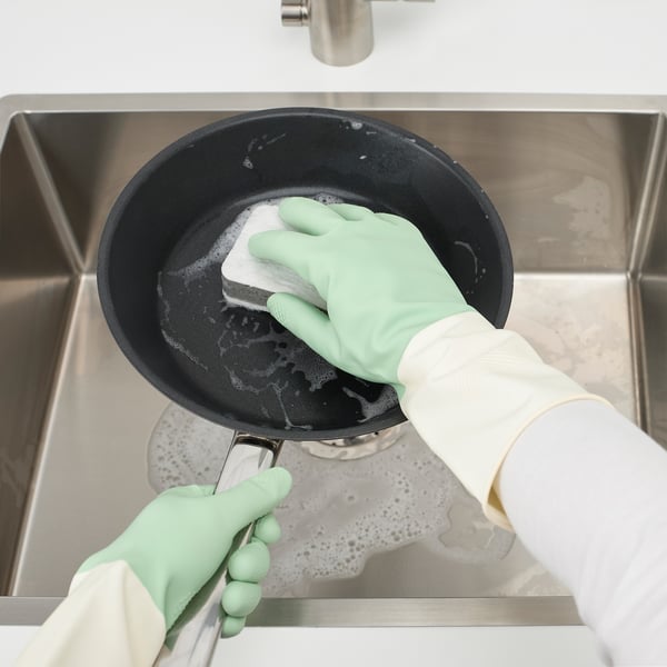 A person wearing green dish gloves washes a black pan in a stainless steel sink, demonstrating kitchen cleaning.