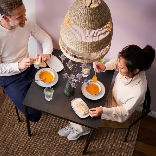 A couple enjoys soup at a black sandberg table with a wicker pendant lamp overhead.