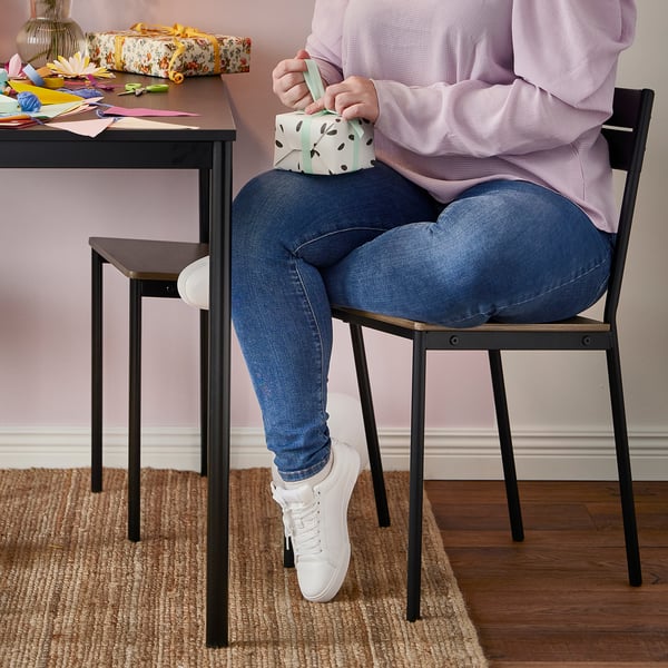 Person wrapping gift box at table with sansberg table and chair, wearing jeans and trainers.
