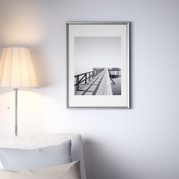 Black and white a wooden dock on a wall. Large silver frame above a bed with a lamp and a pillow.