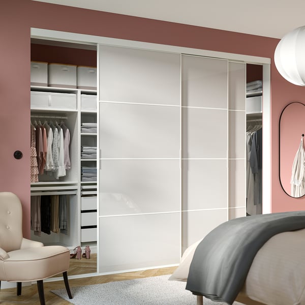 Modern bedroom with large SKYTTA wardrobe, white sliding doors, pink walls, organised clothes, beige chair, grey rug, bed, clock.