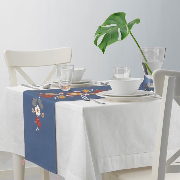 Blue embroidered table runner on white-covered table, surrounded by white chairs and clear glasses.