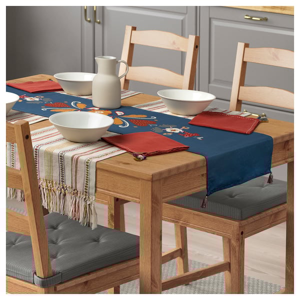 Wooden table set with blue embroidered runner, white bowls, red napkins, and wooden chairs with grey cushions.