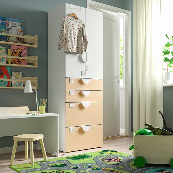 A childs room with a white and beige SMÅSTAD wardrobe, shelves filled with colourful books, a small desk, stool, and a green play mat with toys.