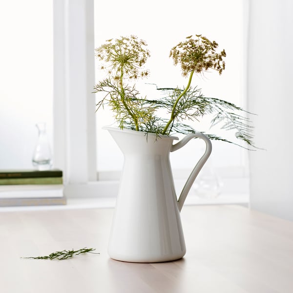 White ceramic jug-shaped vase holding dried flowers, standing on a light-colored surface beside a window, with additional stems on the floor.